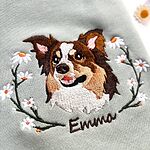 Close-up of the embroidered dog portrait: a brown and white Australian Shepherd face framed by daisy branches, with the name Emma in brown script