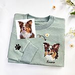 Folded light green sweatshirt with an embroidered dog face and the name Emma under it, no wreath, a brown paw print on the sleeve and the original dog photo beside it
