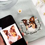 Phone showing the original photo of an Australian Shepherd next to the embroidered portrait of the same dog in a daisy wreath on a sweatshirt