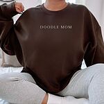 Woman sitting in an oversized Dark Chocolate crewneck sweatshirt embroidered with DOODLE MOM in light serif capitals
