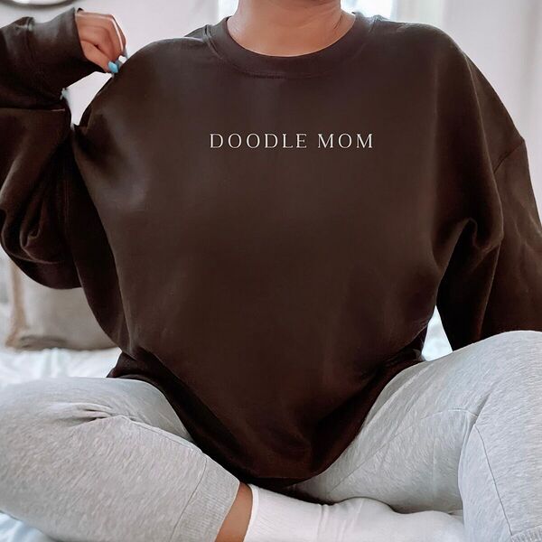 Woman sitting in an oversized Dark Chocolate crewneck sweatshirt embroidered with DOODLE MOM in light serif capitals