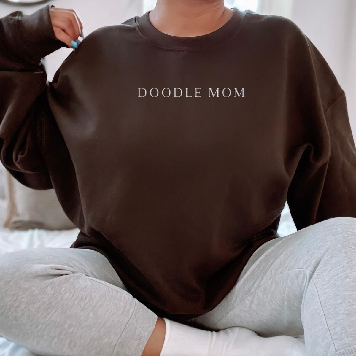 Doodle Mom Crewneck – Dark Chocolate Woman sitting in an oversized Dark Chocolate crewneck sweatshirt embroidered with DOODLE MOM in light serif capitals