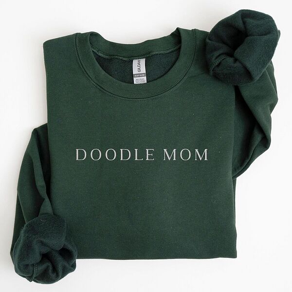 Forest Green crewneck sweatshirt laid flat with a rolled cuff, embroidered with DOODLE MOM in thin serif capitals across the chest