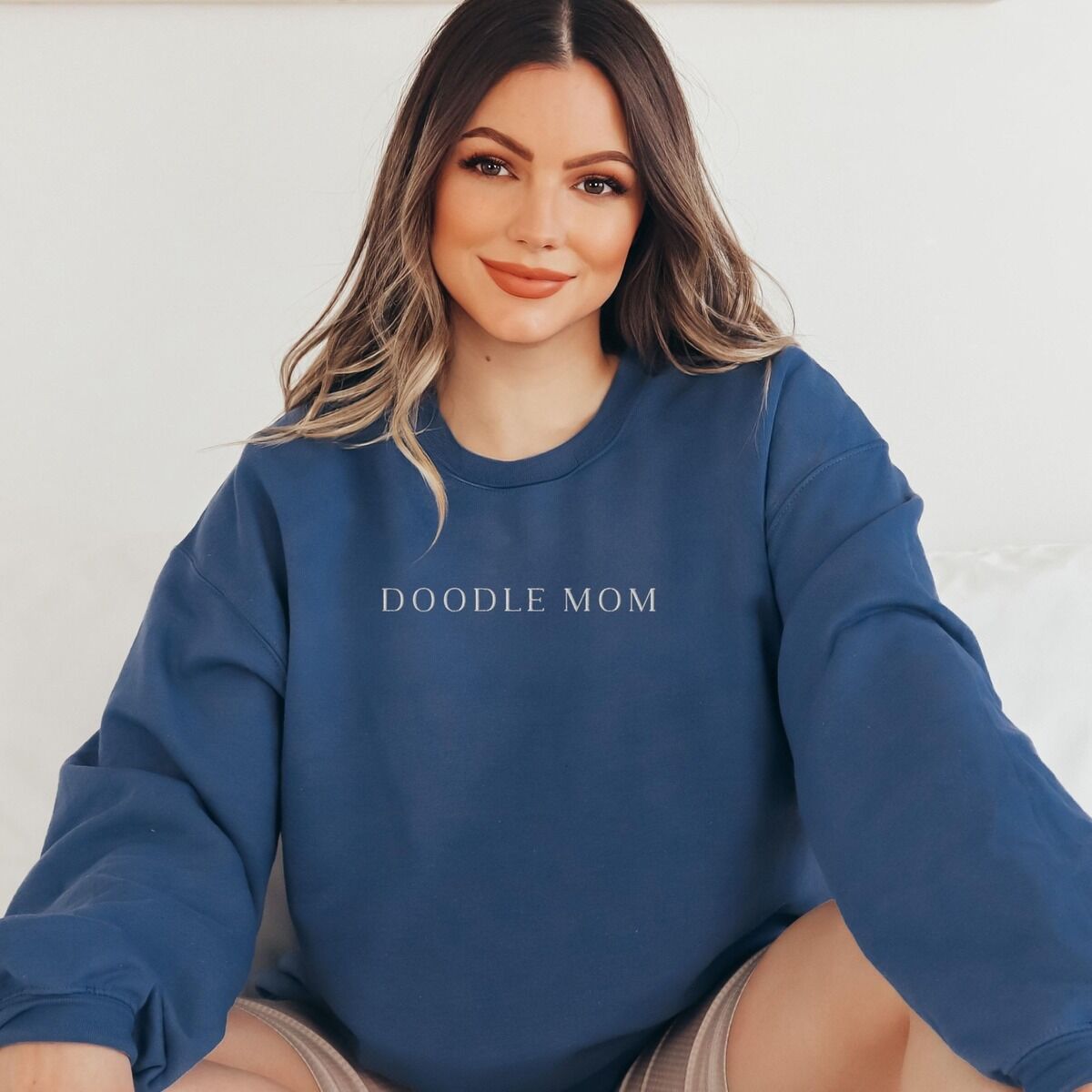 Doodle Mom Crewneck – Indigo Blue Smiling woman in an Indigo Blue crewneck sweatshirt embroidered with DOODLE MOM in light serif capitals across the chest