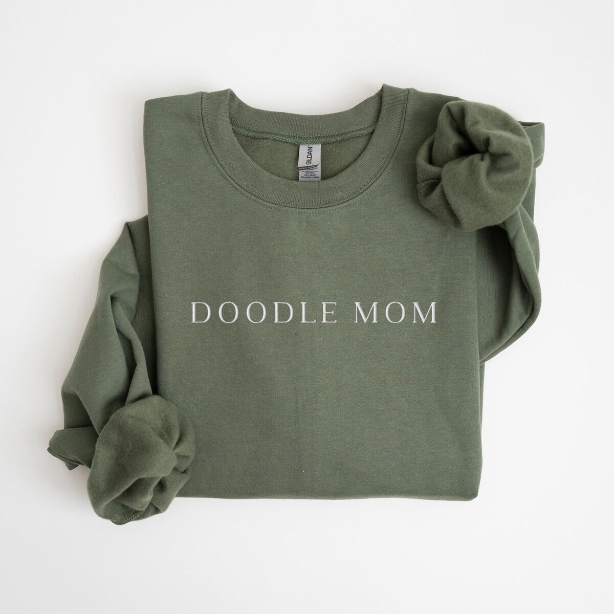 Doodle Mom Crewneck – Military Green Military Green crewneck sweatshirt laid flat with a rolled cuff, embroidered with DOODLE MOM in light serif capitals across the chest