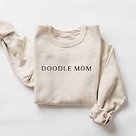 Sand crewneck sweatshirt laid flat with a rolled cuff, embroidered with DOODLE MOM in dark serif capitals across the chest