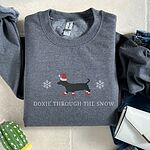 Dark Heather crewneck sweatshirt laid flat beside jeans and a white purse, embroidered with a black dachshund in a Santa hat and red booties between two snowflakes above the words Doxie Through the Snow