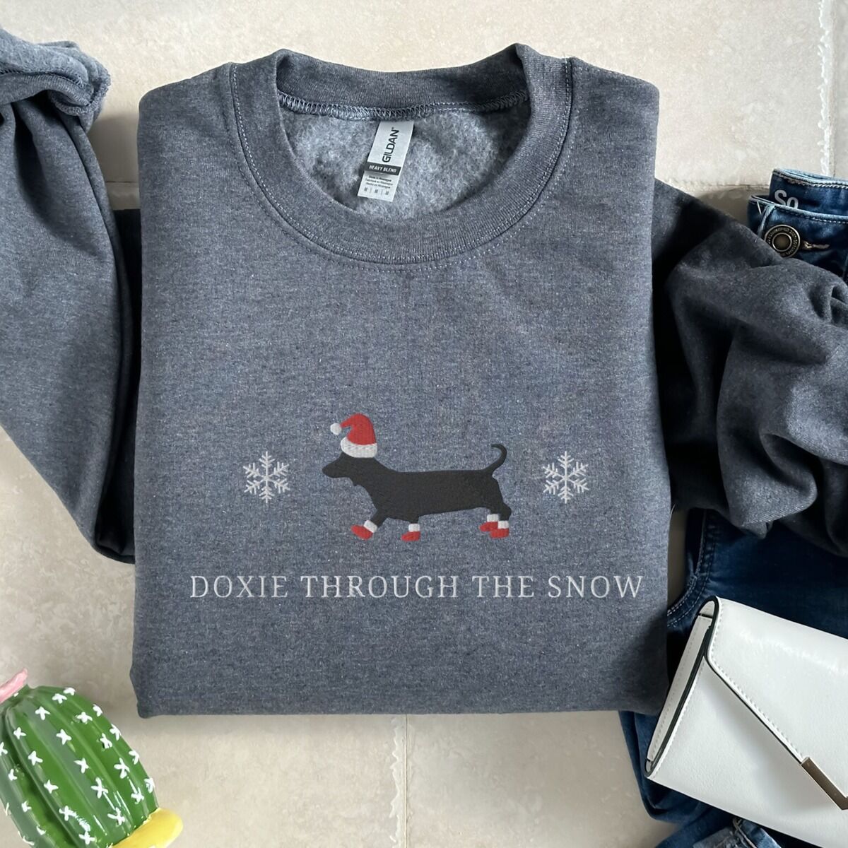 Doxie Through the Snow Sweatshirt – Dark Heather Dark Heather crewneck sweatshirt laid flat beside jeans and a white purse, embroidered with a black dachshund in a Santa hat and red booties between two snowflakes above the words Doxie Through the Snow