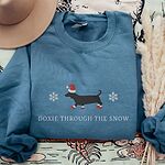 Indigo Blue crewneck sweatshirt laid flat with a felt hat and pampas grass, embroidered with a black dachshund in a Santa hat and red booties between two snowflakes above the words Doxie Through the Snow