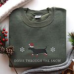 Military Green crewneck sweatshirt laid flat beside pine sprigs, red berries and pinecones, embroidered with a black dachshund in a Santa hat and red booties between two snowflakes above the words Doxie Through the Snow