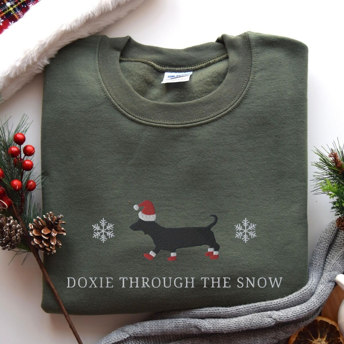 Doxie Through the Snow Sweatshirt – Military Green Military Green crewneck sweatshirt laid flat beside pine sprigs, red berries and pinecones, embroidered with a black dachshund in a Santa hat and red booties between two snowflakes above the words Doxie Through the Snow