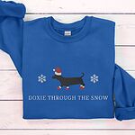 Royal crewneck sweatshirt laid flat on white boards, embroidered with a black dachshund in a Santa hat and red booties between two snowflakes above the words Doxie Through the Snow