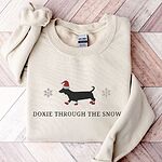 Sand crewneck sweatshirt laid flat on a wood floor, embroidered with a black dachshund in a Santa hat and red booties between two snowflakes above the words Doxie Through the Snow