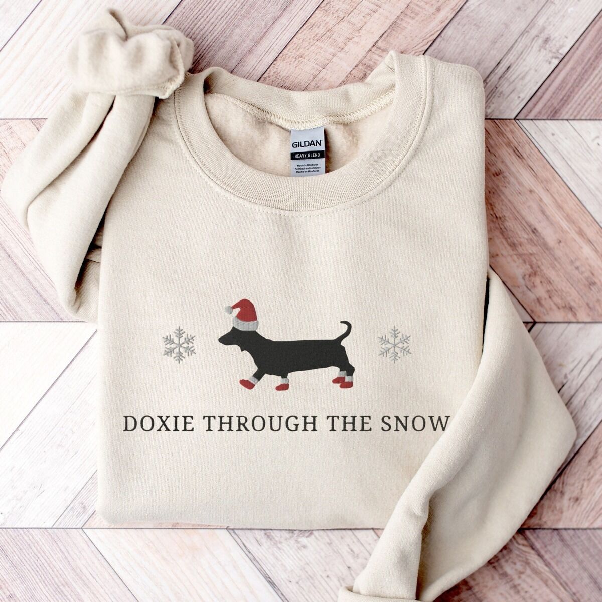 Doxie Through the Snow Sweatshirt – Sand Sand crewneck sweatshirt laid flat on a wood floor, embroidered with a black dachshund in a Santa hat and red booties between two snowflakes above the words Doxie Through the Snow