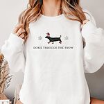 Woman holding the collar of a White crewneck sweatshirt, embroidered with a black dachshund in a Santa hat and red booties between two snowflakes above the words Doxie Through the Snow