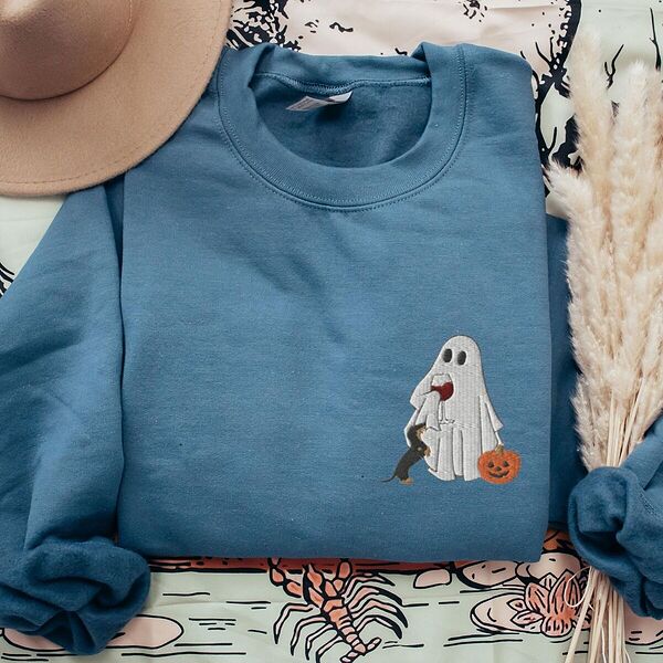 Indigo Blue crewneck sweatshirt laid flat with a brown hat and pampas grass, embroidered on the left chest with a ghost sipping red wine, a black and tan dachshund and a jack-o'-lantern