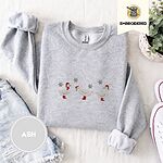 Ash crewneck sweatshirt folded with denim shorts, white sneakers and a potted plant, embroidered with three white ducks in Santa hats and red rain boots, one in a red scarf, among silver snowflakes