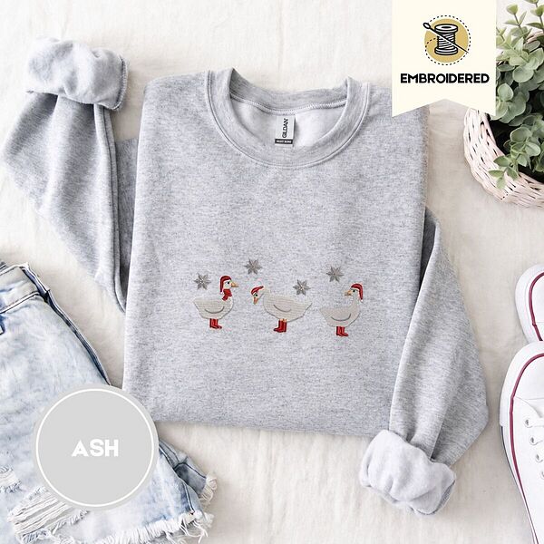 Ash crewneck sweatshirt folded with denim shorts, white sneakers and a potted plant, embroidered with three white ducks in Santa hats and red rain boots, one in a red scarf, among silver snowflakes