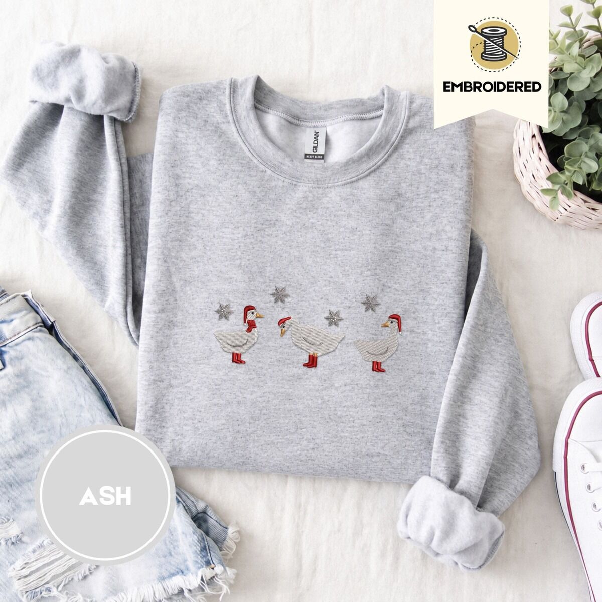 Duck in Boots Sweatshirt – Ash Ash crewneck sweatshirt folded with denim shorts, white sneakers and a potted plant, embroidered with three white ducks in Santa hats and red rain boots, one in a red scarf, among silver snowflakes
