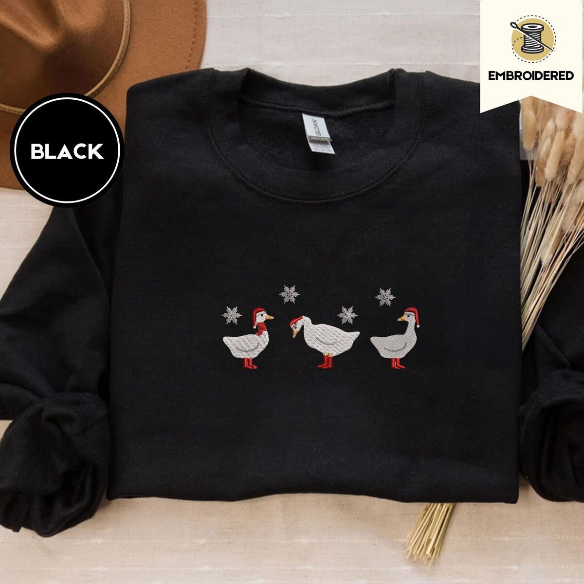 Duck in Boots Sweatshirt – Black Black crewneck sweatshirt with a brown felt hat and dried bunny tail grass, embroidered with three white ducks in Santa hats and red rain boots, one in a red scarf, among silver snowflakes