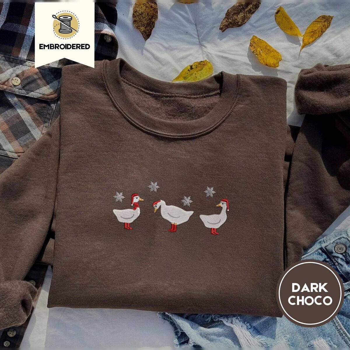 Duck in Boots Sweatshirt – Dark Chocolate Dark Chocolate crewneck sweatshirt on a plaid flannel shirt with yellow autumn leaves and denim shorts, embroidered with three white ducks in Santa hats and red rain boots, one in a red scarf, among silver snowflakes