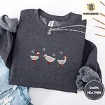 Dark Heather crewneck sweatshirt folded with ripped jeans and a silver chain necklace, embroidered with three white ducks in Santa hats and red rain boots, one in a red scarf, among silver snowflakes