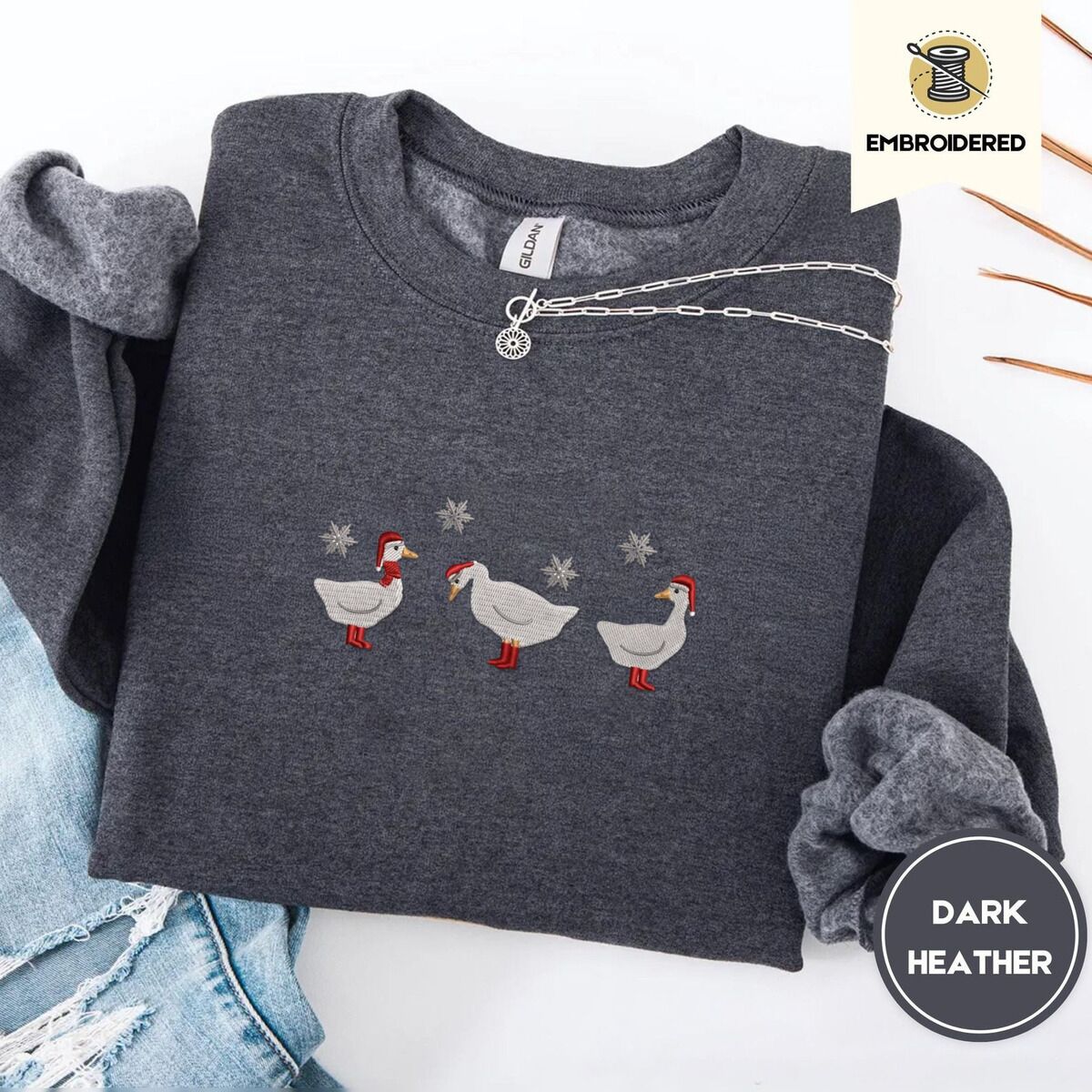 Duck in Boots Sweatshirt – Dark Heather Dark Heather crewneck sweatshirt folded with ripped jeans and a silver chain necklace, embroidered with three white ducks in Santa hats and red rain boots, one in a red scarf, among silver snowflakes