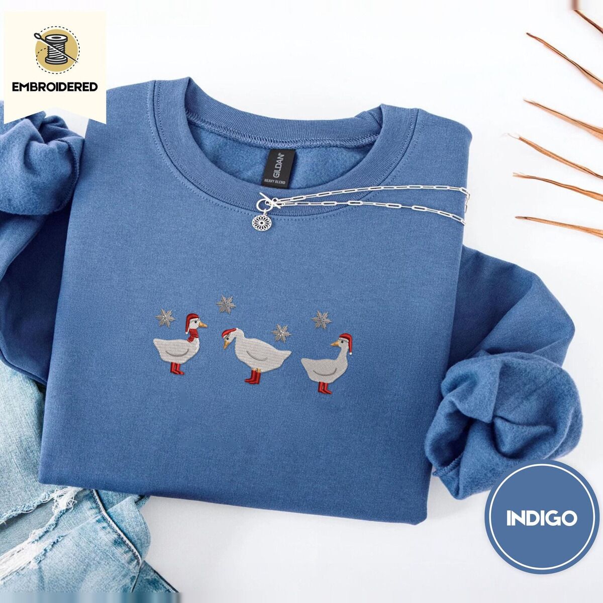 Duck in Boots Sweatshirt – Indigo Blue Indigo Blue crewneck sweatshirt folded with light jeans and dried twigs, embroidered with three white ducks in Santa hats and red rain boots, one in a red scarf, among silver snowflakes