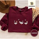 Maroon crewneck sweatshirt with a brown felt hat and dried bunny tail grass, embroidered with three white ducks in Santa hats and red rain boots, one in a red scarf, among silver snowflakes