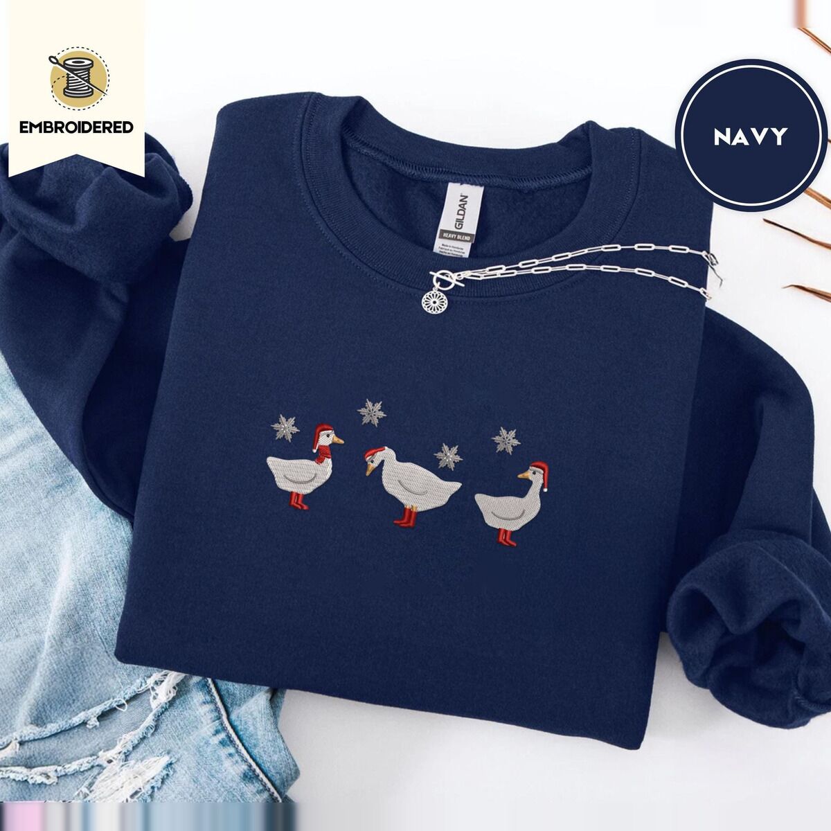 Duck in Boots Sweatshirt – Navy Navy crewneck sweatshirt folded with ripped jeans and a silver chain necklace, embroidered with three white ducks in Santa hats and red rain boots, one in a red scarf, among silver snowflakes