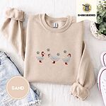 Sand crewneck sweatshirt folded with denim shorts, white sneakers and a potted plant, embroidered with three white ducks in Santa hats and red rain boots, one in a red scarf, among silver snowflakes