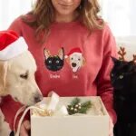Woman holding a gift box in a red sweatshirt embroidered with a black cat in reindeer antlers and a golden retriever in a Santa hat