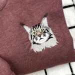 Close-up of an embroidered long-haired tabby cat face with yellow eyes and white whiskers on a maroon heather sweatshirt