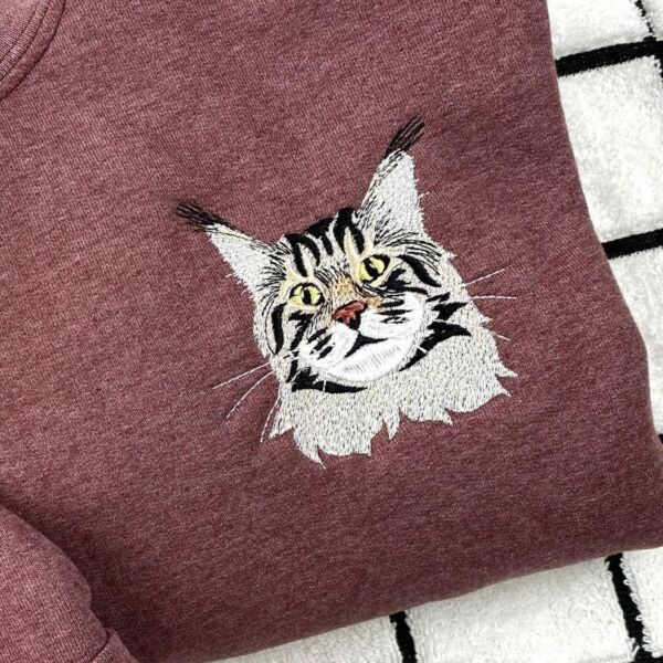 Close-up of an embroidered long-haired tabby cat face with yellow eyes and white whiskers on a maroon heather sweatshirt