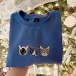 Blue sweatshirt with three embroidered cat faces, a Siamese, a tuxedo and a tabby, in front of a Christmas tree