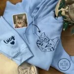 Light blue embroidered cat hoodie with a line art tabby cat face on the left chest and the name Lucy with a heart on the sleeve, next to the cat's photo
