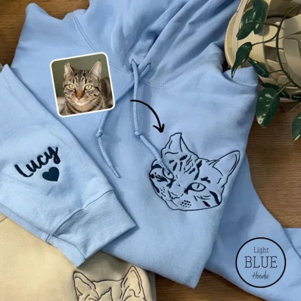 Light blue embroidered cat hoodie with a line art tabby cat face on the left chest and the name Lucy with a heart on the sleeve, next to the cat's photo