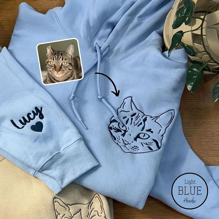 Light blue embroidered cat hoodie with a line art tabby cat face on the left chest and the name Lucy with a heart on the sleeve, next to the cat's photo