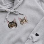 Two embroidered tabby cat portraits on the chest of a grey hoodie, with a small black heart stitched on the sleeve