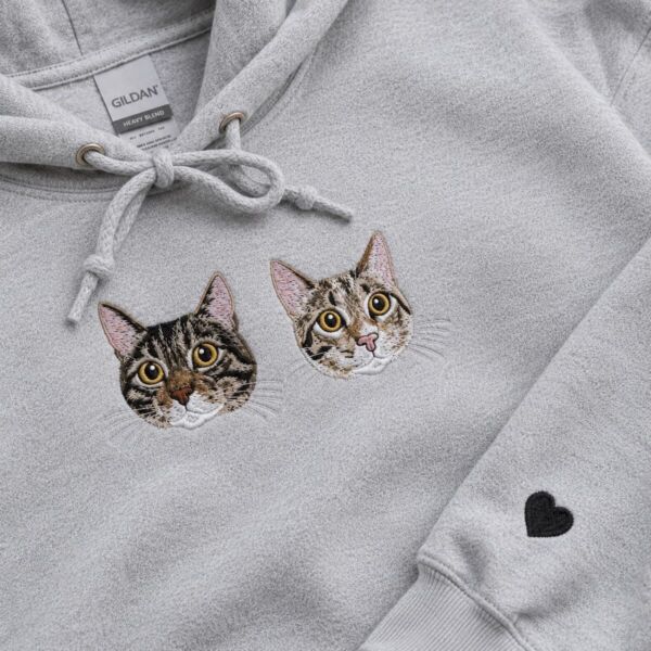 Two embroidered tabby cat portraits on the chest of a grey hoodie, with a small black heart stitched on the sleeve