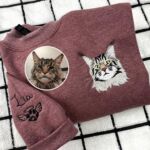 Maroon heather embroidered cat sweatshirt with a long-haired tabby cat portrait on the chest and the name Lia with an angel paw on the sleeve, beside the original photo