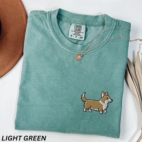 Light Green Comfort Colors T-shirt embroidered with a cute cartoon corgi in golden tan and white trotting on the left chest
