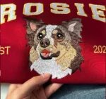 Close-up of an embroidered merle Australian shepherd face with brown eyes and an open mouth on a red sweatshirt