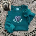 Folded forest green crewneck with the embroidered dog portrait, daisy wreath and the name Gus, shown next to the dog's photo