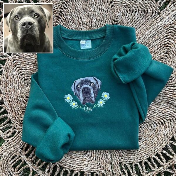 Folded forest green crewneck with the embroidered dog portrait, daisy wreath and the name Gus, shown next to the dog's photo