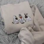 Embroidered ghost dogs Halloween sweatshirt folded on a gray rug, with orange and black pumpkin buckets