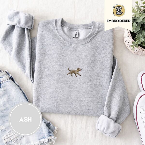 Ash crewneck sweatshirt folded with denim shorts, white sneakers and a potted plant, embroidered with a mini apricot goldendoodle trotting in a gold collar with its tongue out, in the center of the chest