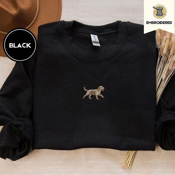 Black crewneck sweatshirt with a brown felt hat and dried bunny tail grass, embroidered with a mini apricot goldendoodle trotting in a gold collar with its tongue out, in the center of the chest