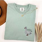 Bay T-shirt laid flat with a necklace and a dried palm leaf, embroidered with a little white goose with the word silly arched over its back on the left chest