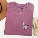 Berry T-shirt laid flat with a necklace and a dried palm leaf, embroidered with a little white goose with the word silly arched over its back on the left chest